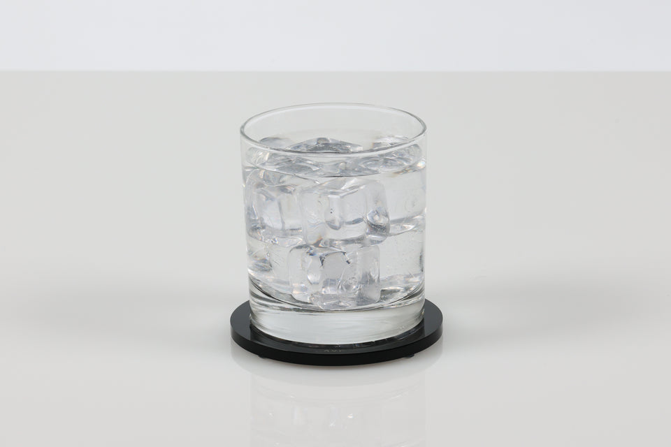 Clear glass of water with ice cubes on a black round lucite coaster on a white background