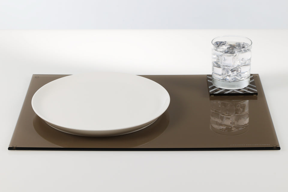 table setting, bronze rectangle placemat. on top of the placemat sits a white circular plate and a square bronze chevron pattern coater with a glass of water on it. 