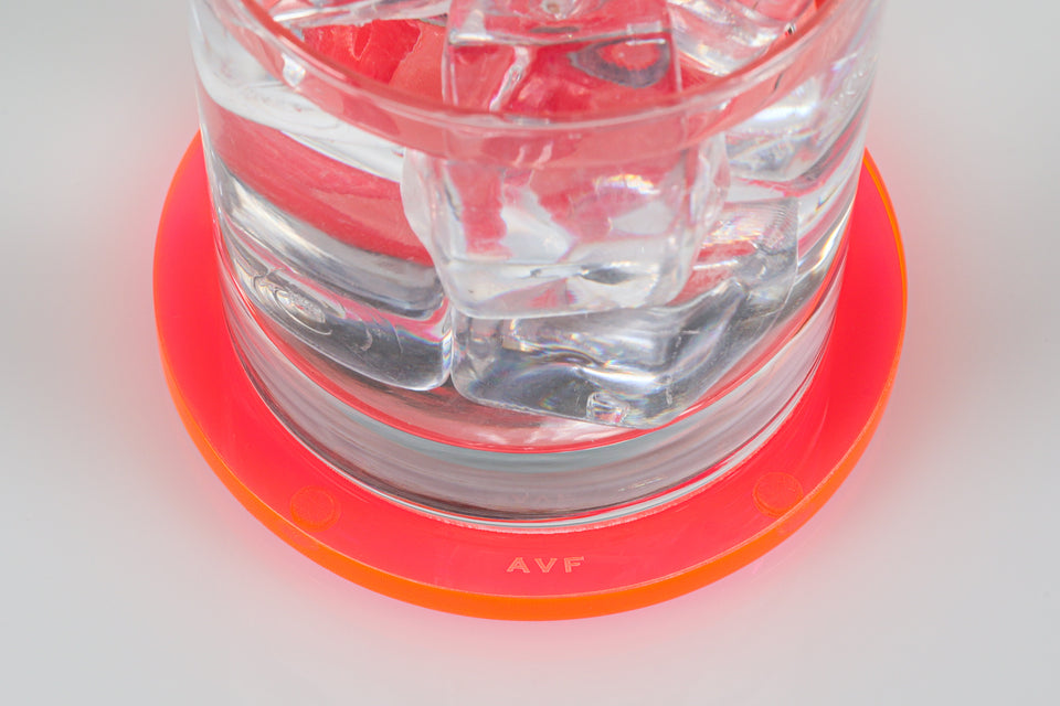 Clear glass with ice cubes on a Pink bullseye coaster on a white surface. focused on the AVF logo. 