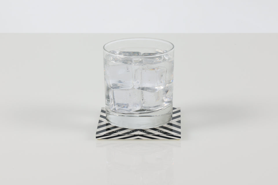 Clear glass with water on a black and white X-Print coaster against a white background. 